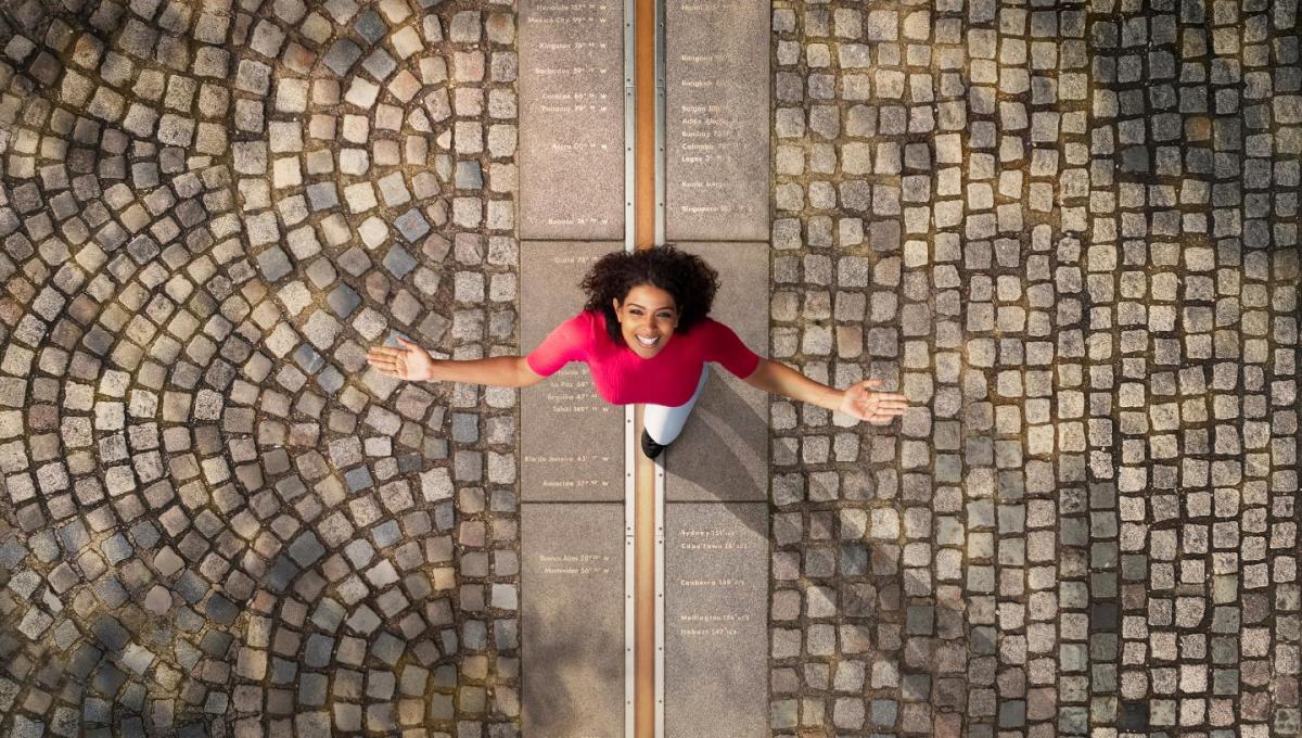 A woman stands on the Prime Meridian line at Royal Observatory Greenwich, looking up and smiling