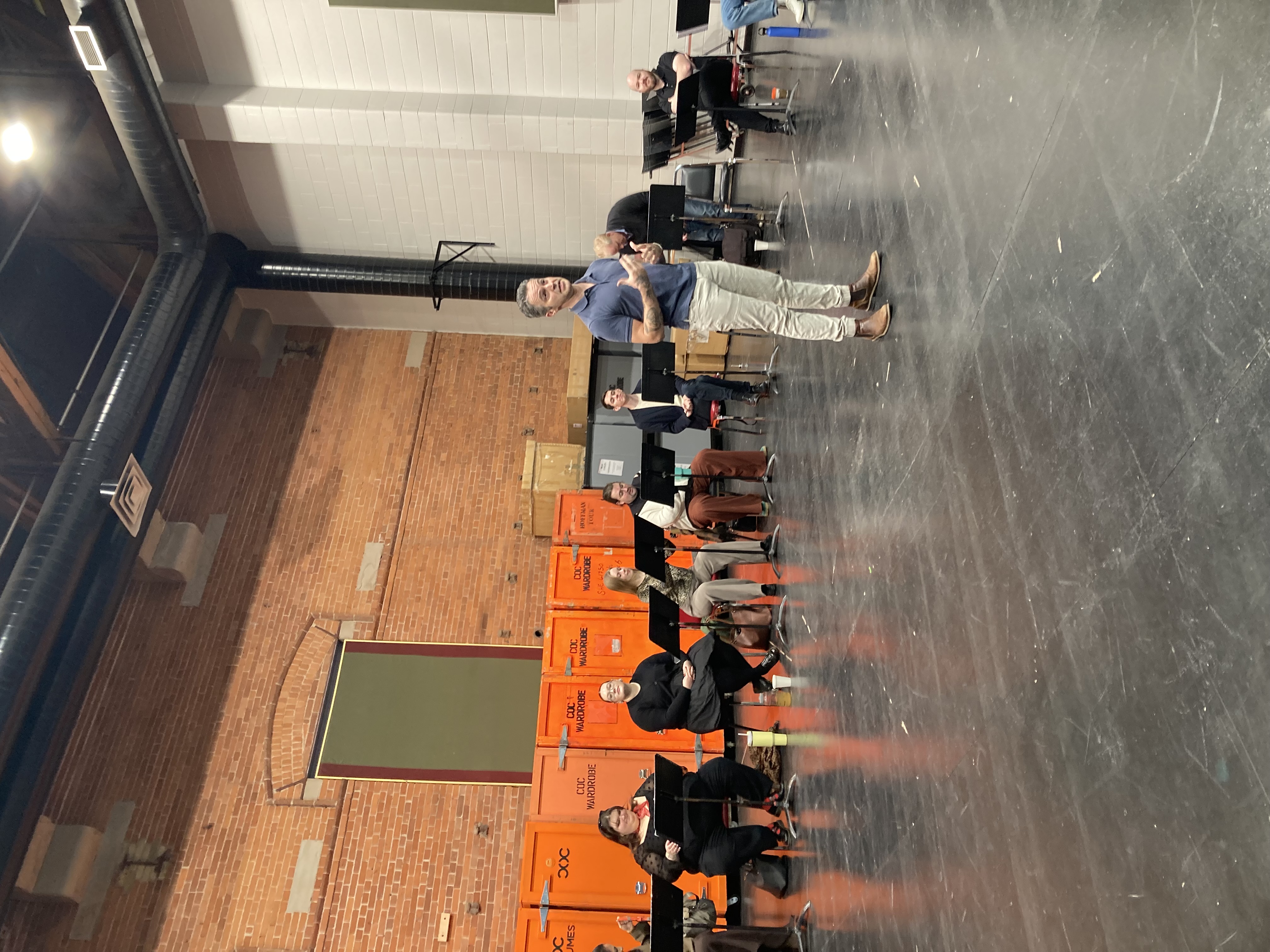 Ian Cusson stands in a large rehearsal room, gesturing towards the unseen audience. He is standing in front of a group of animated opera performers.