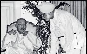 This is a famous black-and-white photo from 1991. It shows the two men who changed India's economy. On the left, Prime Minister P.V. Narasimha Rao is sitting in a large chair wearing a white traditional outfit. On the right, Finance Minister Manmohan Singh is leaning toward him and holding a folder with important papers. They both look very serious because they were making big decisions to open up India to the rest of the world.
