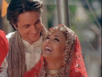 This photo shows a close up of the two main characters, William Darcy and Lalita Bakshi. Lalita is dressed in a beautiful red wedding outfit with a lot of gold jewelry and a sparkling headpiece. She is laughing and looking up at Darcy. Darcy is wearing a cream colored traditional Indian shirt and a tan scarf, and he is looking down at her with a sweet smile. It is a very personal moment that shows how their different worlds eventually came together.