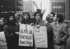 A photo of protest taking during the U.S. Sexual Revolution of the 1960s. It is a black and white photo of a group of men holding two signs. The one of the left of the image states, "I am a sexual being." while the one on the right is too blurry to fully read.