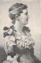 L.T. Meade, the first editor of Atalanta. In this image, she is portrayed in half-body portrait, from head to waist, in a Victorian dress. She is facing almost 90 degrees to the left. Her hair is neatly brushed with small bun. An earring is shown on her right ear.