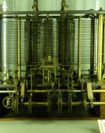 A bronze and gold machine. The inner workings are exposed in columns of bronze and connected with long threads of metal across the top. In between these columns of bronze are columns of metal that house the mathematical units to run calculations. Beneath are a series of gears that move the middle columns. 