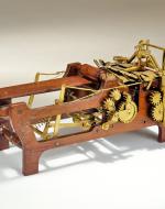 A wooden framed machine shaped like a cradle. The front of the machine is shaped like a printer tray and has very little below it. The back is a mess of gears and levers in bronze affixed to a more solid wooden chassis.