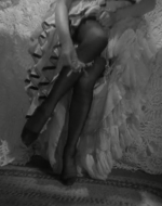 Black and white image of a woman sitting on a bed, taking off her stockings. 