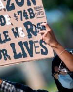 A masked woman holds a sign reading "Amazon made 74 billion while we risked our lives"
