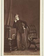 Full length photographic portrait of Prince Leopold standing facing front. His right elbow leans on top of a trolley, his right hand at his chin. He holds his cane in his left hand. Chair to right, on which his cap.