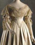 A modern photograph of Victoria's wedding dress. Cream satin, with lace adorning the collar and the sleeves. The bodice is fitted and tapers to a point. The skirt is full.