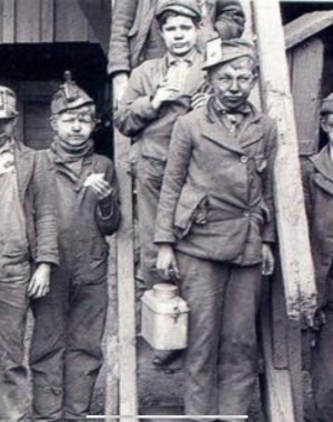 A black and white image of six young boys covered in dust and dirt. Somber expressions cover their faces. Outfits resemble those of chimney sweeps.