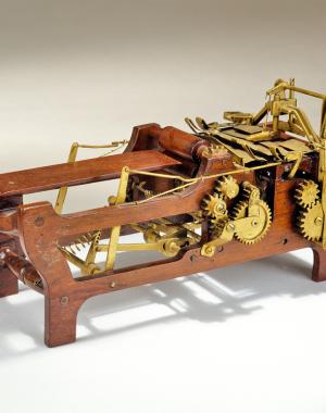A wooden framed machine shaped like a cradle. The front of the machine is shaped like a printer tray and has very little below it. The back is a mess of gears and levers in bronze affixed to a more solid wooden chassis.
