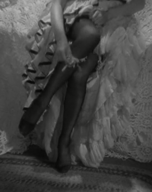 Black and white image of a woman sitting on a bed, taking off her stockings. 