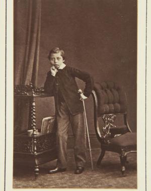 Full length photographic portrait of Prince Leopold standing facing front. His right elbow leans on top of a trolley, his right hand at his chin. He holds his cane in his left hand. Chair to right, on which his cap.