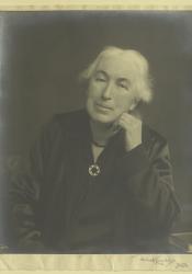 Photograph of Clemence Housman in old age, with white hair, a black dress and brooch, and a dark dress. Her chin rests on her left hand and she is looking directly at the camera. 
