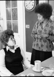 Toni Morrison and Alice Walker are seen in a kitchen. Toni is sitting to the left looking up at Alice, standing to the right.