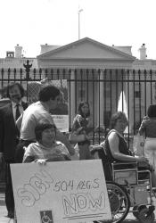 A group of protestors, many are wheelchair users, with Judy Heumann in the forefront holding a handwritten sign that says "Sign 504 regs NOW."