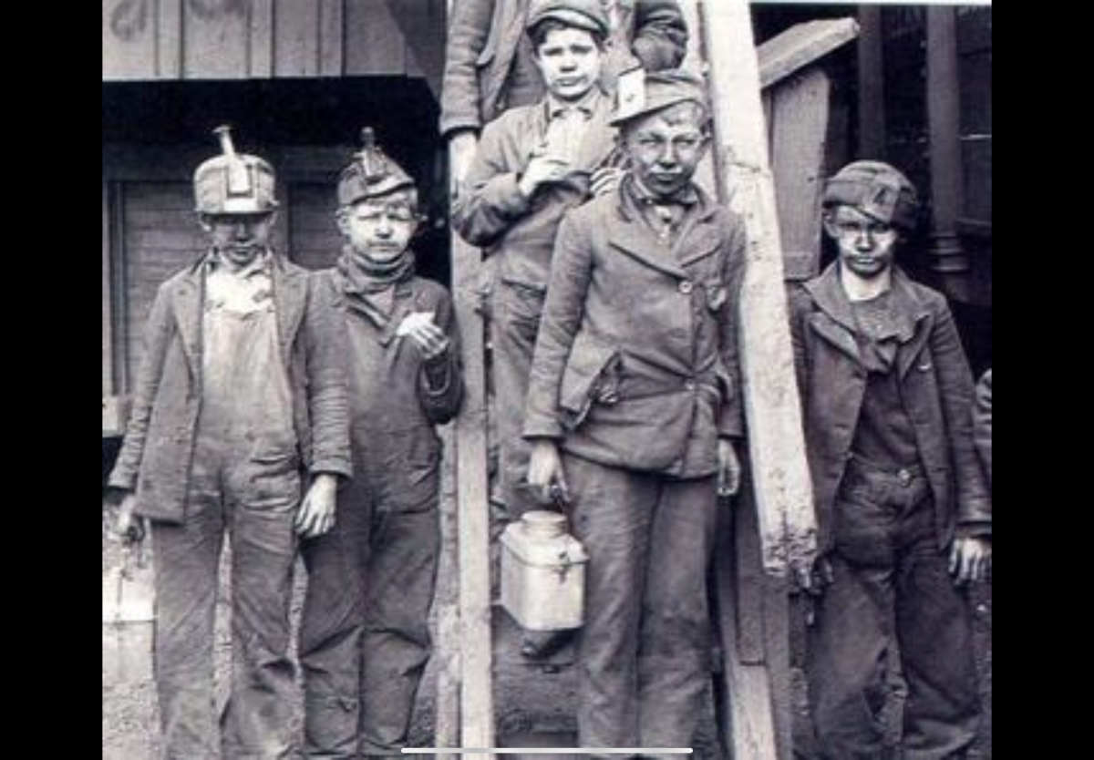 A black and white image of six young boys covered in dust and dirt. Somber expressions cover their faces. Outfits resemble those of chimney sweeps.