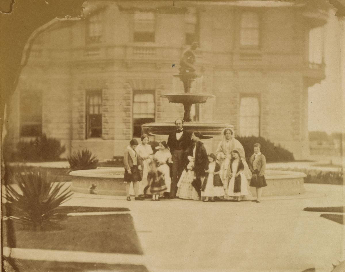 Photograph of a group portrait of Queen Victoria and Prince Albert with their seven eldest children and Queen Victoria's mother, the Duchess of Kent. The group are posed in front of a fountain at Osborne House. The group stand, positioned from left to right are: Albert Edward, Prince of Wales, Princess Victoria, Princess Royal, Prince Arthur, Princess Alice, Prince Albert, Queen Victoria, Princess Louise, the Duchess of Kent, Princess Helena and Prince Alfred.
