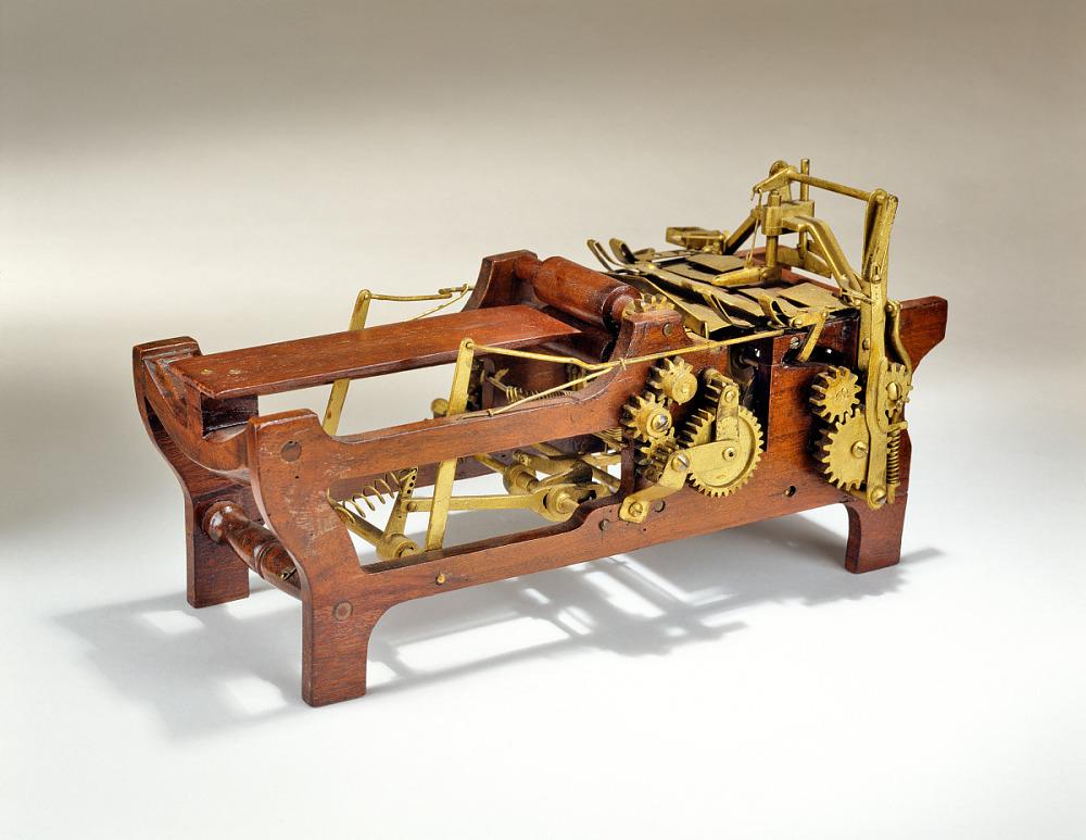 A wooden framed machine shaped like a cradle. The front of the machine is shaped like a printer tray and has very little below it. The back is a mess of gears and levers in bronze affixed to a more solid wooden chassis.