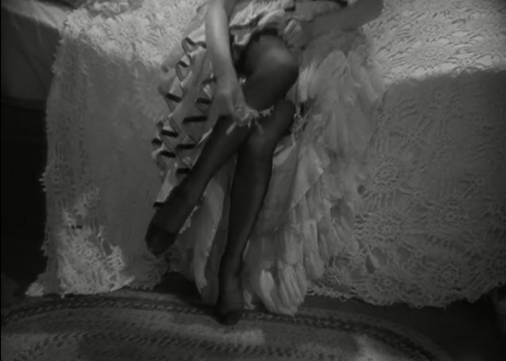 Black and white image of a woman sitting on a bed, taking off her stockings. 