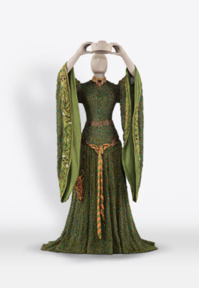A long green dress, with large sleeves and a gold belt. The dress, a Lady Macbeth costume made with beetles’ wings, is on a mannequin that is holding a crown above its head.