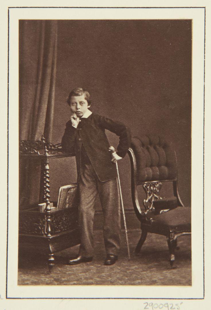 Full length photographic portrait of Prince Leopold standing facing front. His right elbow leans on top of a trolley, his right hand at his chin. He holds his cane in his left hand. Chair to right, on which his cap.